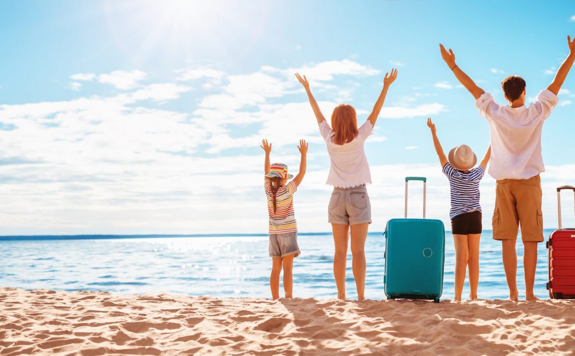 Mother and father with their children standing on the beach with suitcases.